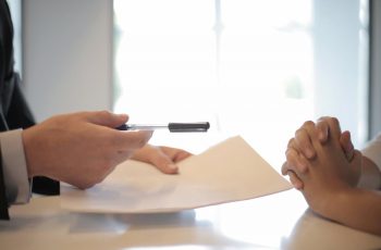 Imagem de um homem oferecendo uma folha e papel para outra pessoa assinar em uma mesa
