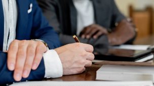 Imagem de um homem assinando documentos do lado de uma pessoa em uma mesa de madeira