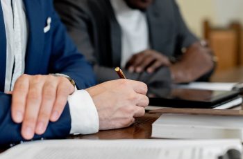 Imagem de um homem assinando documentos do lado de uma pessoa em uma mesa de madeira